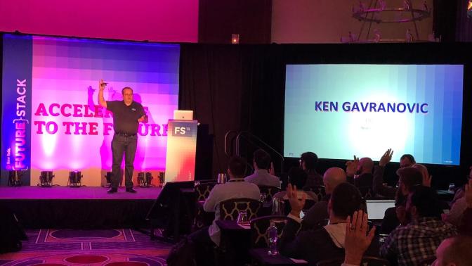 Ken Gavranovic presenting on a stage with his name on the screen as the audience raises hands